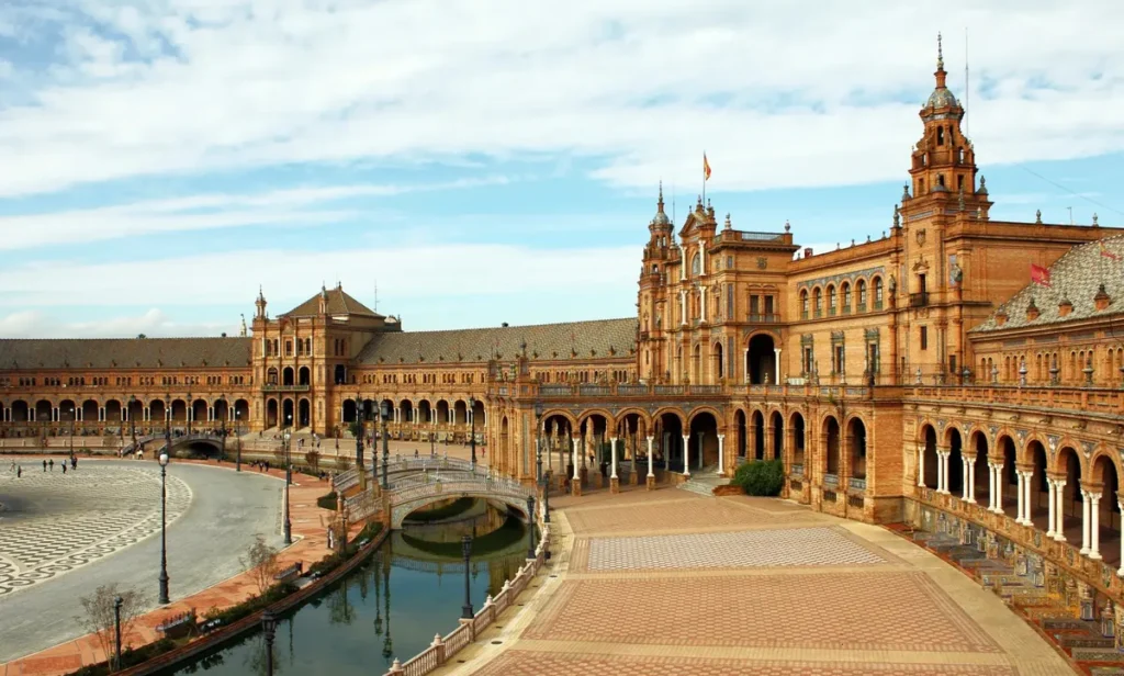 Vista da Plaza de España, um dos lugares mais emblemáticos de Sevilha e uma das obras arquitetônicas mais impressionantes da Andaluzia. Construída para a Exposição Ibero-Americana de 1929, a praça apresenta um conjunto monumental de edifícios semicirculares, torres ornamentadas, galerias de arcos e detalhes em cerâmica colorida. A fotografia destaca uma das pontes que atravessam o canal da praça, além da arquitetura de tijolos avermelhados e dos amplos espaços abertos. Localizada no Parque de María Luisa, a Plaza de España combina influências renascentistas, barrocas e mudéjares, sendo uma parada indispensável em qualquer roteiro turístico por Sevilha.