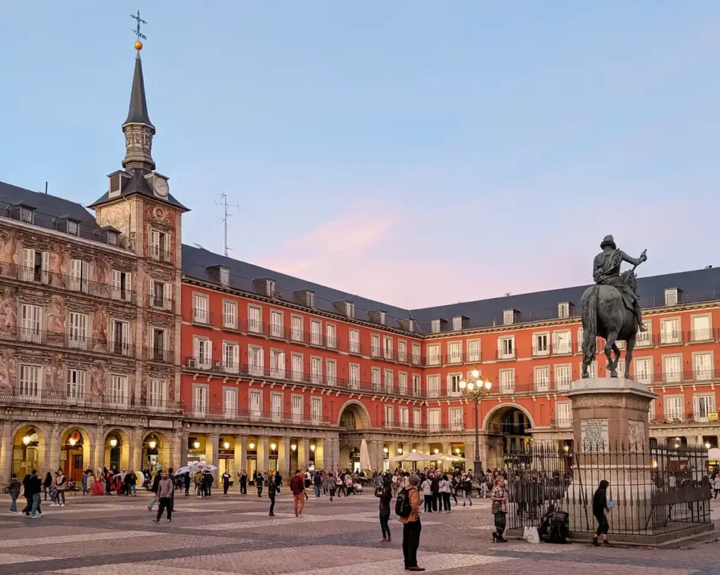 A Plaza Mayor é uma das praças históricas mais famosas de Madrid e um dos principais pontos turísticos do centro da capital espanhola. A fotografia mostra a estátua equestre do rei Filipe III em primeiro plano, cercada pelos tradicionais edifícios de fachadas avermelhadas, janelas simétricas e galerias com arcadas. À esquerda, destaca-se a Casa de la Panadería, reconhecida por sua fachada decorada com pinturas e torres com pináculos. O movimento de turistas, a iluminação dos postes e o céu suave do entardecer revelam a atmosfera cultural e arquitetônica desta praça histórica, construída durante o reinado de Filipe III.