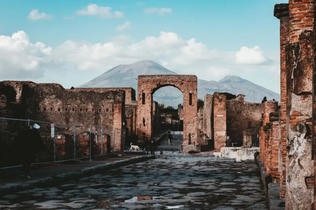 Ruínas da antiga cidade romana de Pompeia, próxima a Nápoles, com uma rua de pedras preservada, construções arqueológicas e o Monte Vesúvio ao fundo. Soterrada pela erupção vulcânica de 79 d.C., Pompeia é um dos sítios arqueológicos mais importantes da Itália e Patrimônio Mundial da UNESCO.