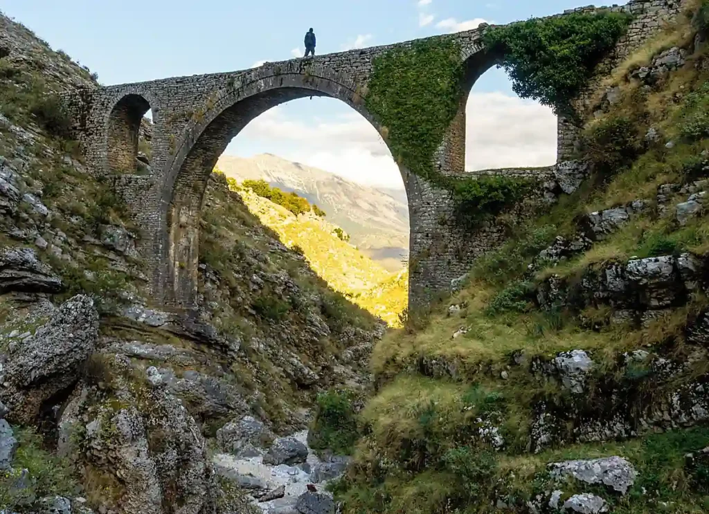 Ponte histórica de pedra na Albânia, exemplo da arquitetura tradicional presente nas antigas rotas comerciais dos Bálcãs. A construção em arcos destaca-se em meio a um vale montanhoso, com vegetação e formações rochosas. As pontes otomanas fazem parte do patrimônio arquitetônico e cultural albanês, refletindo a história das comunidades que atravessavam rios e regiões montanhosas do país.