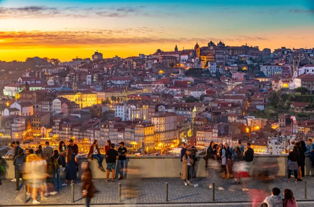 O centro histórico do Porto revela uma das paisagens urbanas mais características de Portugal, com edifícios centenários, igrejas barrocas, ruas em diferentes níveis e um conjunto de telhados avermelhados que acompanha as encostas próximas ao rio Douro. A fotografia registra a cidade durante o pôr do sol, vista de um miradouro movimentado por turistas, com o céu em tons intensos de laranja, amarelo e azul. A iluminação urbana começa a destacar as fachadas históricas, enquanto visitantes observam a paisagem da Ribeira e das colinas do Porto.