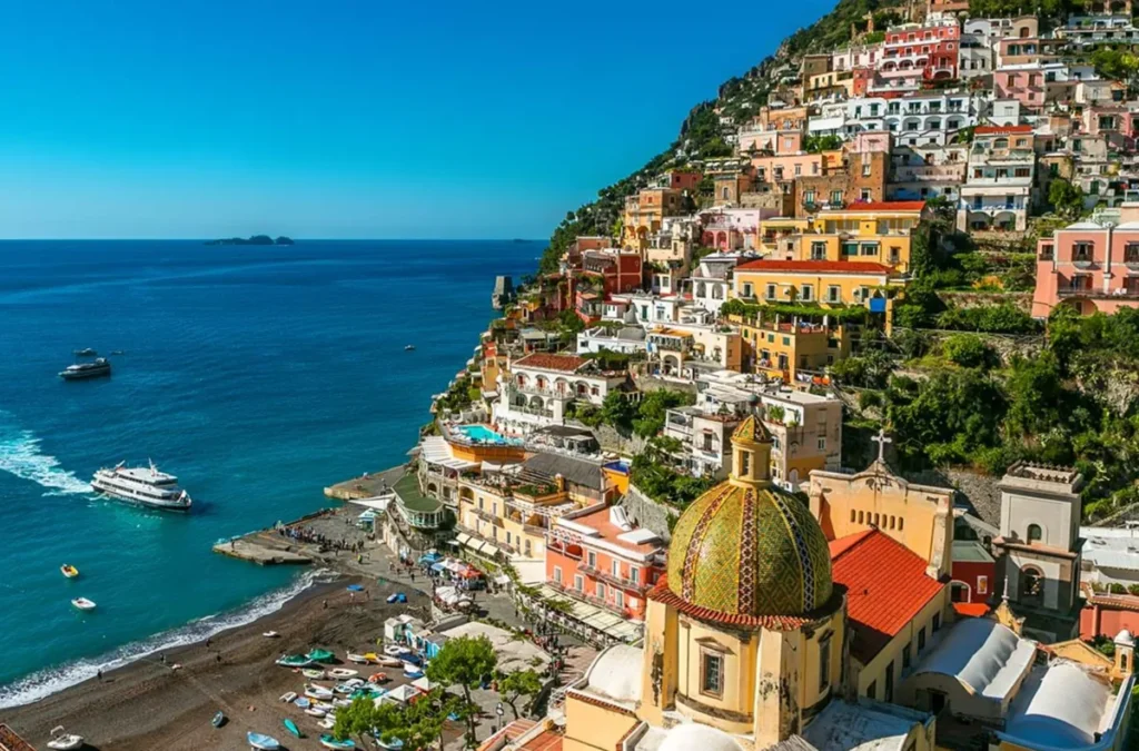Positano é uma das cidades mais fotografadas da Costa Amalfitana, famosa por suas casas coloridas construídas em terraços sobre as encostas íngremes do litoral italiano. A fotografia apresenta uma vista elevada da cidade, destacando a cúpula revestida de azulejos amarelos e verdes da Igreja de Santa Maria Assunta, um dos principais símbolos arquitetônicos de Positano. Abaixo, a praia de Spiaggia Grande reúne pequenas embarcações, visitantes e estruturas turísticas junto ao mar Tirreno. As fachadas em tons de rosa, amarelo, branco e terracota contrastam com o azul profundo do Mediterrâneo, formando uma das paisagens mais reconhecidas do turismo na Itália.