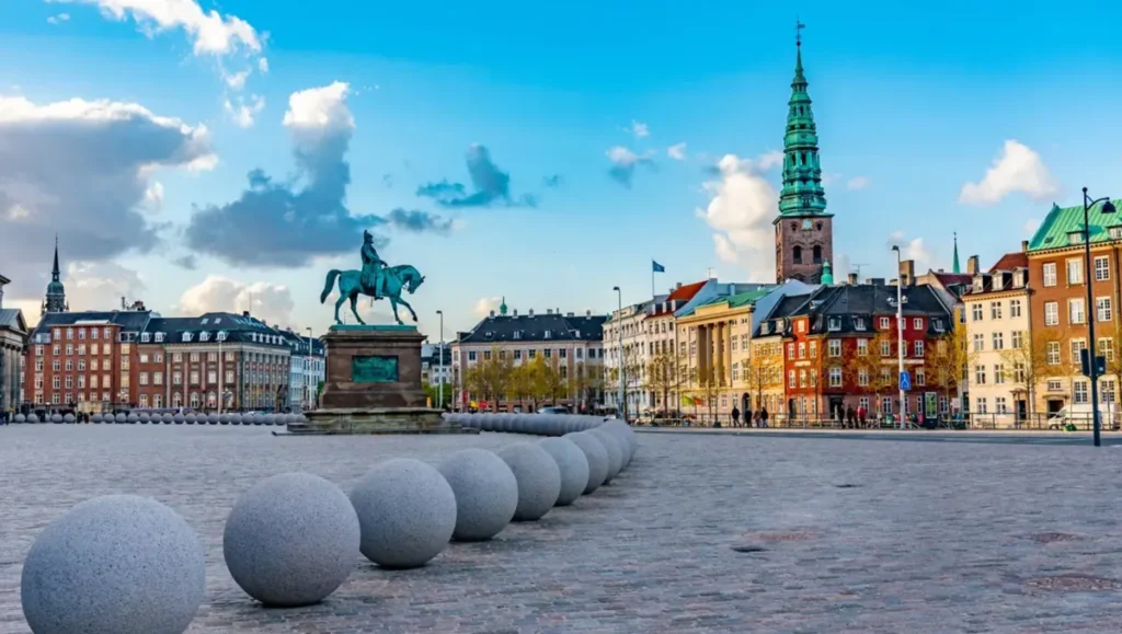 Christiansborg Slotsplads, praça histórica de Copenhague, localizada diante do Palácio de Christiansborg, sede do Parlamento dinamarquês. O monumento equestre de Frederico VII e os edifícios históricos compõem uma paisagem urbana característica da capital da Dinamarca, conhecida por sua arquitetura, história monárquica e patrimônio cultural.