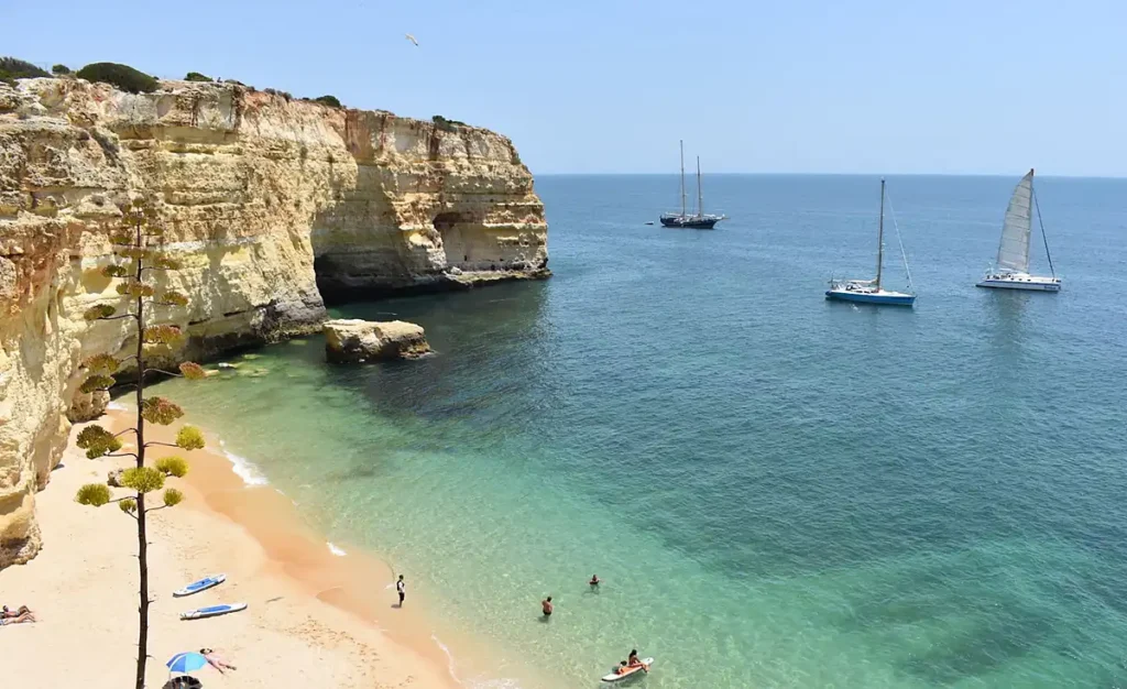 Paisagem do litoral do Algarve, no sul de Portugal, com uma pequena praia de areia dourada protegida por falésias calcárias, mar transparente e veleiros próximos à costa. A região é conhecida por suas enseadas, praias escondidas entre rochas, passeios de barco, stand-up paddle e paisagens naturais que atraem visitantes durante os meses mais quentes do ano.