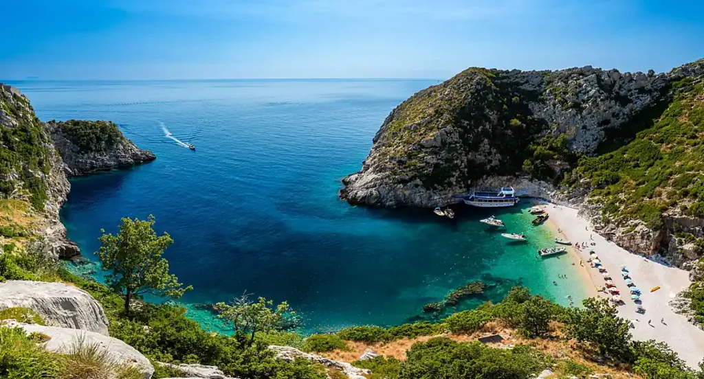 Praia de águas cristalinas na Riviera Albanesa, região costeira do sul da Albânia banhada pelo Mar Jônico. A paisagem apresenta uma enseada protegida por falésias, vegetação mediterrânea e águas transparentes em diferentes tons de azul. O litoral albanês é conhecido por suas praias naturais, passeios de barco, pequenas baías e cenários que fazem da região um destino cada vez mais procurado para viagens de verão pelo Mediterrâneo.