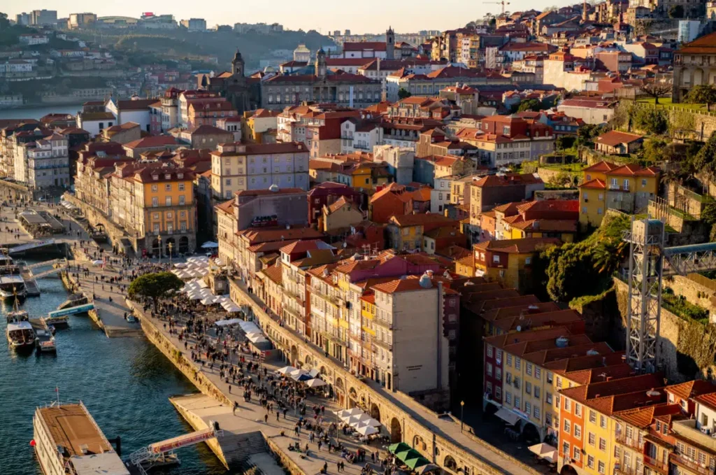 A Ribeira do Porto é um dos bairros mais emblemáticos da cidade do Porto, no norte de Portugal, e integra o centro histórico classificado como Patrimônio Mundial pela UNESCO. A fotografia apresenta uma vista elevada do Cais da Ribeira, às margens do rio Douro, destacando os edifícios históricos de fachadas estreitas e coloridas, telhados de terracota, restaurantes com mesas ao ar livre e o movimento de visitantes no passeio ribeirinho. A luz quente do fim da tarde ilumina as construções tradicionais e revela a paisagem urbana marcada por ruas estreitas, igrejas históricas e encostas densamente ocupadas.