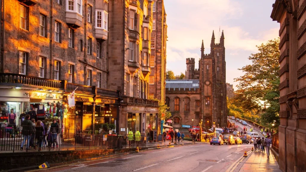 Vista de uma rua histórica no centro de Edimburgo, capital da Escócia, durante o entardecer. A fotografia destaca a arquitetura tradicional escocesa, com edifícios de pedra de vários andares, fachadas antigas, vitrines de lojas e estabelecimentos comerciais iluminados pela luz dourada do sol. Ao longo da rua, pedestres e veículos circulam entre construções históricas, enquanto uma imponente torre de estilo neogótico se destaca ao fundo. A paisagem urbana representa o charme da Old Town de Edimburgo, região reconhecida por suas ruas históricas, arquitetura medieval e atmosfera cultural. A Royal Mile e seus arredores estão entre os principais atrativos turísticos da cidade.
