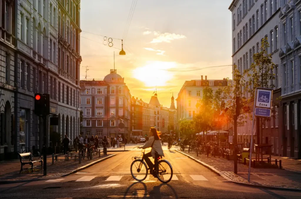 Copenhague, capital da Dinamarca, durante o pôr do sol, com ciclistas circulando pelas ruas históricas da cidade. Reconhecida mundialmente por sua infraestrutura cicloviária e mobilidade sustentável, Copenhagen combina arquitetura escandinava, edifícios tradicionais e uma cultura urbana em que a bicicleta é um dos principais meios de transporte.