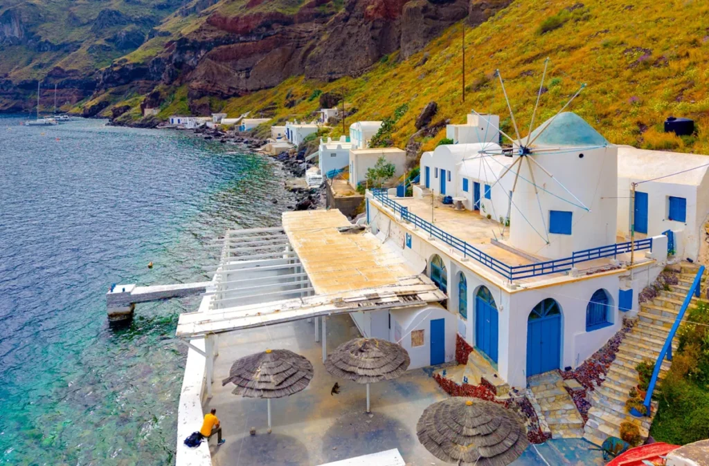 Paisagem litorânea de Santorini, uma das ilhas mais famosas das Cíclades, na Grécia, conhecida por suas construções brancas, detalhes arquitetônicos azuis e impressionantes formações vulcânicas. A fotografia mostra um pequeno conjunto de edifícios tradicionais à beira do mar Egeu, com paredes caiadas de branco, portas e janelas azuis, terraços junto à água e um moinho de vento característico da arquitetura grega. Ao fundo, encostas rochosas com vegetação verde e amarelada contrastam com as águas transparentes da costa. A composição destaca o ambiente mediterrâneo, a arquitetura tradicional das ilhas gregas e a paisagem natural de Santorini.