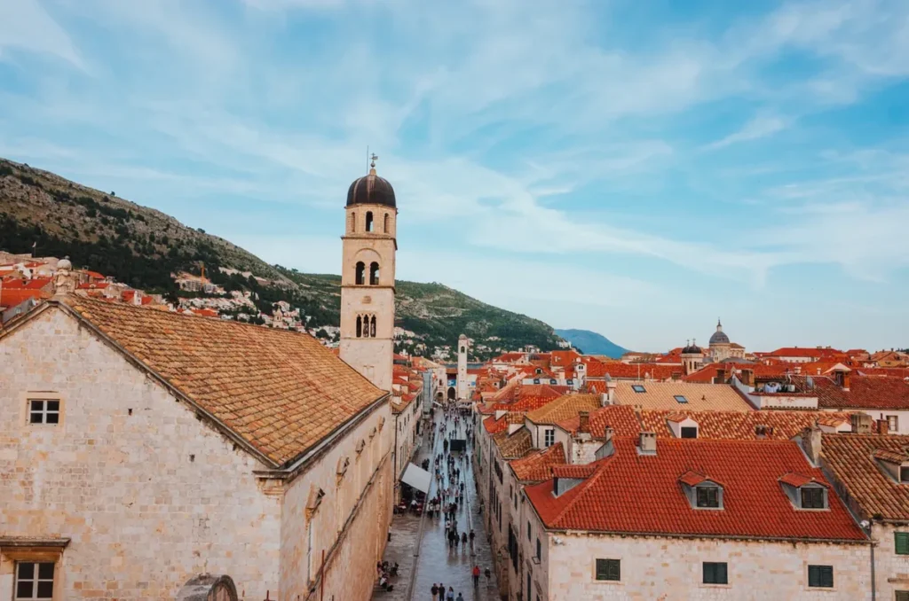 Vista elevada da Stradun, também conhecida como Placa, a principal rua do centro histórico de Dubrovnik, na Croácia. A fotografia mostra a tradicional via pavimentada em pedra calcária, cercada por edifícios históricos de fachadas claras e telhados avermelhados. À esquerda, destaca-se a torre sineira do Mosteiro Franciscano, enquanto visitantes caminham pela movimentada rua que atravessa a Cidade Antiga. Ao fundo, as montanhas e construções distribuídas pelas encostas completam a paisagem. A Stradun é um dos pontos turísticos mais conhecidos de Dubrovnik e concentra importantes atrações históricas, culturais e arquitetônicas da cidade.