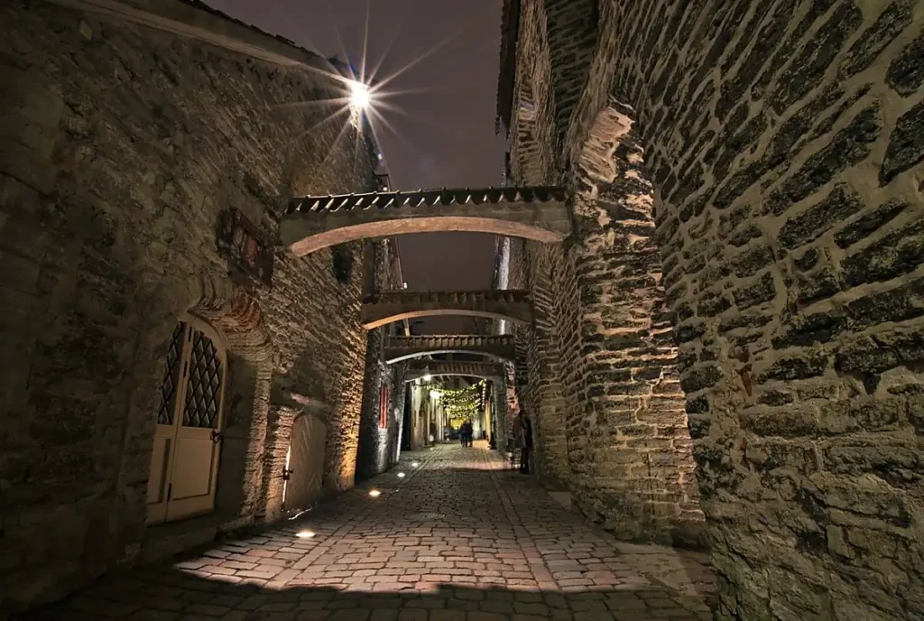 Rua medieval iluminada à noite no centro histórico de Tallinn, na Estônia. As construções de pedra, os arcos que atravessam a passagem e o calçamento antigo mostram a atmosfera histórica das vielas da cidade.