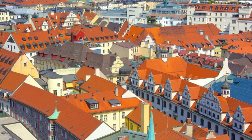 Vista panorâmica dos telhados e edifícios históricos do centro de Munique, capital da Baviera, no sul da Alemanha. A fotografia revela uma paisagem urbana marcada por telhados de cerâmica avermelhada, fachadas ornamentadas, pequenas torres e construções que refletem diferentes períodos da arquitetura europeia. Entre edifícios residenciais, igrejas e antigas construções comerciais, o centro histórico de Munique preserva características tradicionais da cidade, mesmo após as extensas reconstruções realizadas depois da Segunda Guerra Mundial. A imagem destaca a diversidade arquitetônica da capital bávara e o contraste entre suas construções históricas e os edifícios mais modernos.