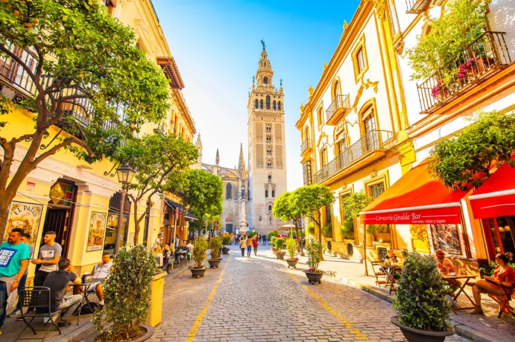 Vista da Torre Giralda, um dos principais cartões-postais de Sevilha, destacando-se ao fundo de uma charmosa rua do centro histórico da cidade. A fotografia mostra o ambiente típico da Andaluzia, com edifícios de fachadas claras, varandas de ferro trabalhado, laranjeiras e restaurantes com mesas ao ar livre. Originalmente construída como minarete durante o período islâmico, a Giralda foi posteriormente adaptada como campanário da Catedral de Sevilha, tornando-se um dos monumentos mais representativos da arquitetura histórica espanhola. A paisagem combina o patrimônio cultural da cidade com a atmosfera animada de suas ruas, cafés e estabelecimentos tradicionais.