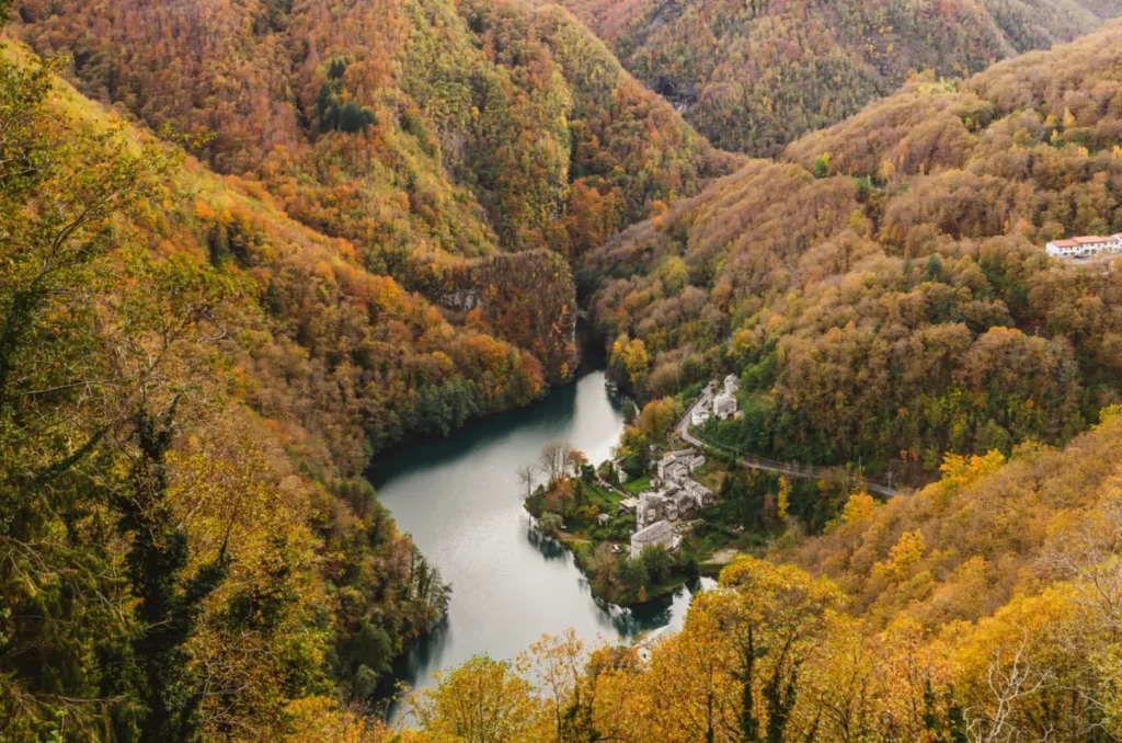Paisagem de outono com um lago cercado por montanhas, florestas douradas e um pequeno vilarejo à beira da água.