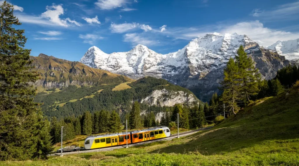 Trem de montanha amarelo e laranja circulando por uma ferrovia panorâmica na região de Jungfrau, nos Alpes Berneses, Suíça. A composição destaca extensos campos verdes, florestas alpinas e um impressionante conjunto de montanhas cobertas de neve, incluindo os famosos picos Eiger, Mönch e Jungfrau. O cenário representa uma das experiências mais características do turismo suíço: percorrer ferrovias alpinas cercadas por paisagens naturais de grande beleza, próximas a destinos como Grindelwald, Wengen e Kleine Scheidegg.