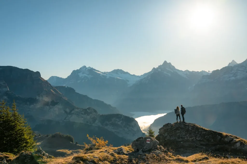 Dois visitantes contemplam uma paisagem montanhosa dos Alpes Suíços a partir de um mirante natural localizado sobre uma formação rochosa. O panorama apresenta uma sucessão de picos alpinos, montanhas parcialmente cobertas de neve, encostas florestadas e um vale preenchido por nuvens baixas. A luz suave do sol ilumina as montanhas e destaca a grandiosidade do cenário. A fotografia representa a experiência de caminhadas e trilhas panorâmicas na região de Interlaken e Jungfrau, reconhecida por suas paisagens de alta montanha e atividades de turismo de natureza.