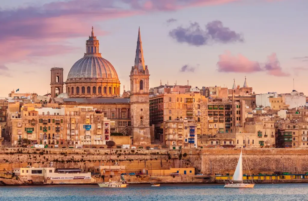 Valletta ao pôr do sol, com a cúpula da Basílica de Nossa Senhora do Carmo e a torre da Catedral Anglicana de São Paulo destacando-se sobre a arquitetura histórica da capital de Malta. A luz dourada ilumina os edifícios de pedra calcária e cria uma das vistas mais emblemáticas da cidade mediterrânea.