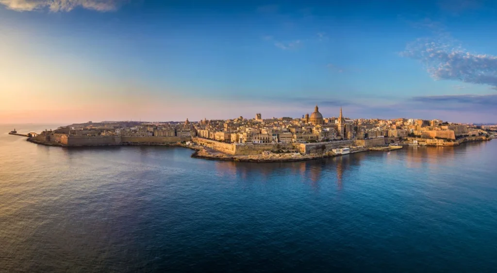 Vista panorâmica de Valletta, capital de Malta, ao entardecer, destacando as muralhas históricas, igrejas barrocas e construções de pedra calcária. Situada em uma península entre dois portos naturais, a cidade é reconhecida pela UNESCO e preserva um dos conjuntos arquitetônicos mais importantes do Mediterrâneo.