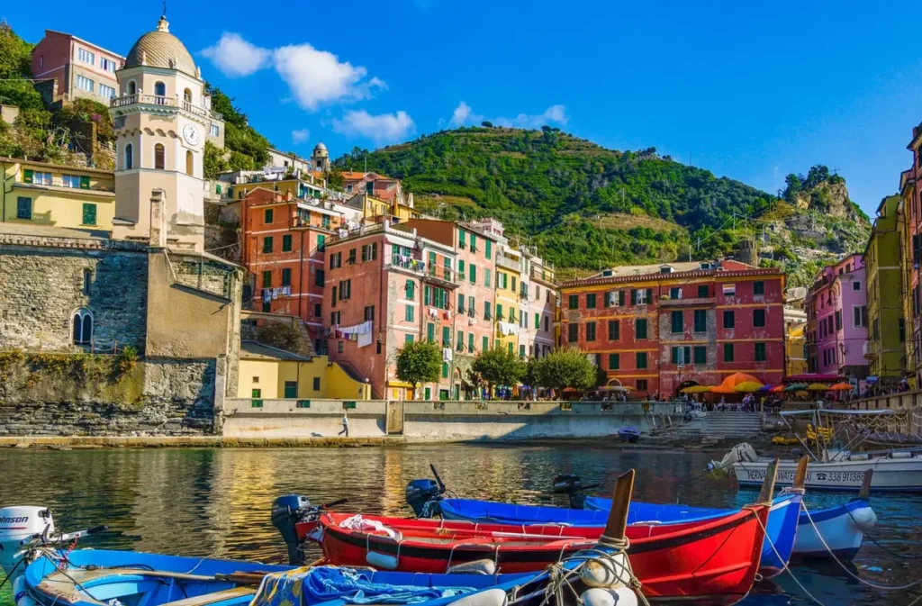 Vista do charmoso porto de Vernazza, uma das vilas mais famosas de Cinque Terre, na Itália. A fotografia apresenta pequenas embarcações de pesca coloridas flutuando nas águas tranquilas do Mar da Ligúria, com destaque para a Igreja de Santa Margherita di Antiochia, cuja torre histórica se eleva entre as construções tradicionais. Ao fundo, casas em tons de rosa, amarelo e vermelho acompanham o relevo montanhoso, formando uma paisagem característica da Riviera Italiana. O porto de Vernazza é um dos principais pontos turísticos da vila, oferecendo acesso a restaurantes, cafés, pequenas praias e passeios pelo centro histórico. A combinação entre arquitetura medieval, cultura marítima e beleza natural transforma Vernazza em um dos destinos mais fotografados de Cinque Terre.