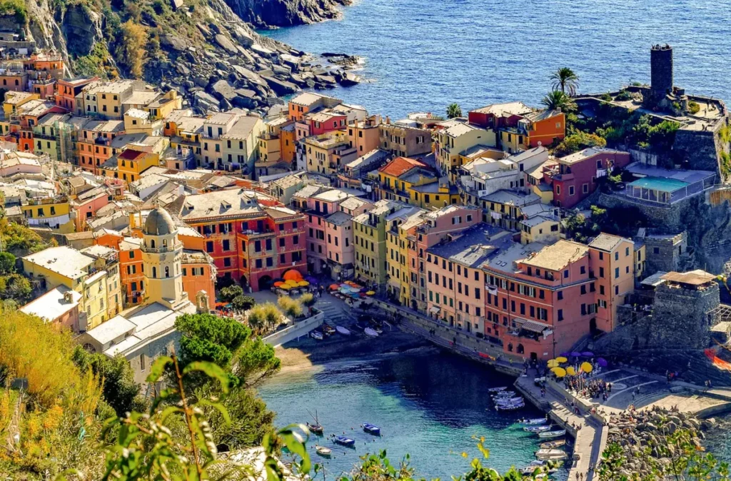 Vista aérea da vila de Vernazza, localizada no Parque Nacional de Cinque Terre, na região da Ligúria, Itália. A imagem revela o conjunto de casas coloridas distribuídas entre as encostas rochosas e o litoral mediterrâneo, com destaque para a Igreja de Santa Margherita di Antiochia, o Castelo Doria e a pequena enseada que abriga o porto da vila. As construções históricas em tons de amarelo, rosa e vermelho contrastam com as águas azuis do Mar da Ligúria, criando uma das paisagens mais emblemáticas da Riviera Italiana. Vernazza é conhecida por sua arquitetura tradicional, ruas estreitas, trilhas costeiras e restaurantes especializados em frutos do mar e pratos típicos da Ligúria. A vila integra o conjunto de Cinque Terre, reconhecido internacionalmente por sua importância histórica, cultural e paisagística.