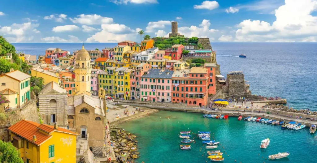 Vista panorâmica de Vernazza, uma das cinco vilas que compõem Cinque Terre, na região da Ligúria, no litoral noroeste da Itália. A fotografia destaca as tradicionais casas coloridas construídas sobre as encostas, a Igreja de Santa Margherita di Antiochia, o pequeno porto de águas azul-turquesa e a torre do Castelo Doria, que domina a paisagem. Cercada pelo Mar da Ligúria, Vernazza é considerada uma das localidades mais encantadoras de Cinque Terre, conhecida por suas ruas estreitas, construções históricas, restaurantes à beira-mar e paisagens mediterrâneas. A vila faz parte da área reconhecida como Patrimônio Mundial pela UNESCO e atrai visitantes interessados em turismo cultural, gastronomia italiana, trilhas panorâmicas e passeios pelas vilas costeiras.