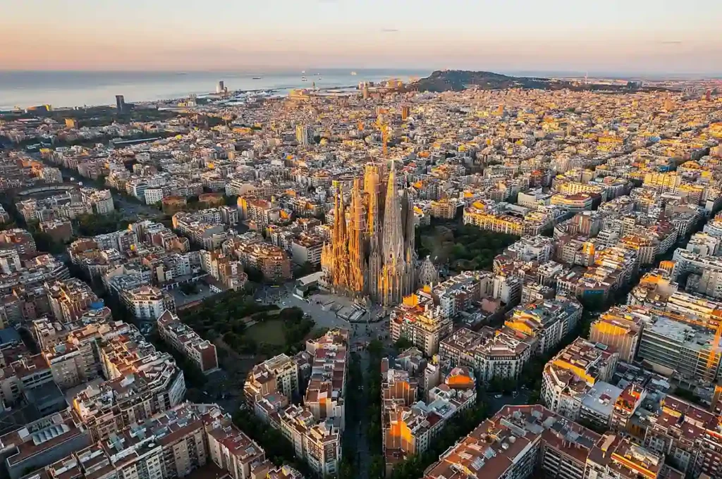 A vista aérea de Barcelona revela a Sagrada Família cercada pela malha urbana do Eixample, famosa por suas quadras geométricas. Ao fundo, o Mar Mediterrâneo completa a paisagem de uma das cidades mais visitadas da Espanha.