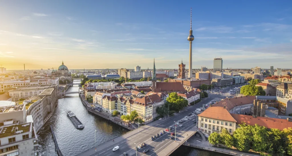 Vista panorâmica do centro de Berlim, na Alemanha, durante o final da tarde, com destaque para a Fernsehturm, a famosa Torre de TV de Berlim, que domina o horizonte da cidade. A fotografia revela o Rio Spree atravessando a paisagem urbana, com pontes, embarcações turísticas e edifícios históricos distribuídos ao longo de suas margens. À esquerda, a cúpula da Catedral de Berlim se destaca entre as construções da Ilha dos Museus, enquanto a arquitetura moderna e as amplas avenidas evidenciam o contraste entre o passado e o presente da capital alemã. A luz dourada do pôr do sol completa a paisagem de uma das cidades mais importantes da Europa.